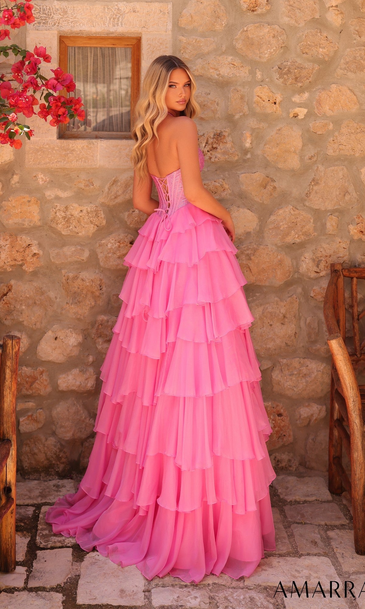 A woman wearing the Long Prom Dress: Amarra 89287—a strapless pink gown with a chiffon tiered A-line skirt—stands by a stone wall outdoors, looking over her shoulder.